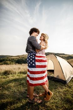 An attractive couple in love wrapped in american flag enjoying in front a tent at the campsite.