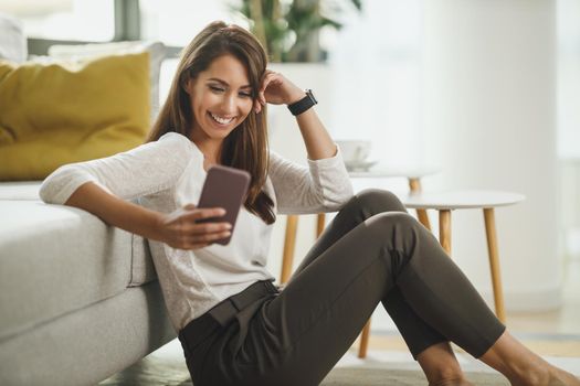 Beautiful, young woman sitting next to the window, having leisure time, holding a smart phone and surfing social media at her home.