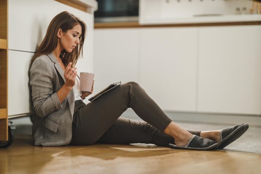 Shot of a young woman drinking coffee and using a digital tablet while sitting on the floor in her kitchen.