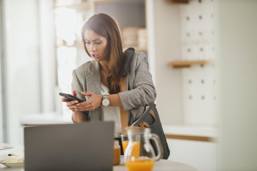 Shot of a multi-tasking young business woman typing on her smartphone in her kitchen while getting ready to go to work.