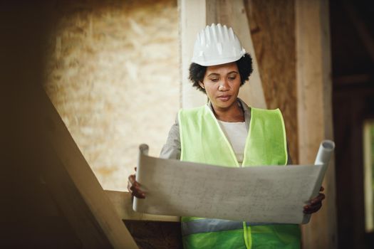 Shot of an African female architect checking plans at the construction site of a new wooden house. She is wearing protective workwear and white helmet.