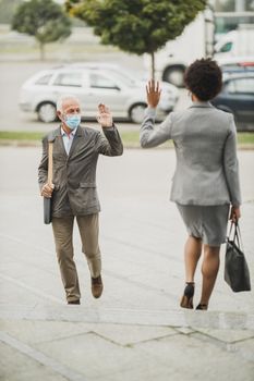 Shot of successful senior businessman and his black female colleague using protective mask and greeting with wave hands during COVID-19 pandemic to avoid handshakes.
