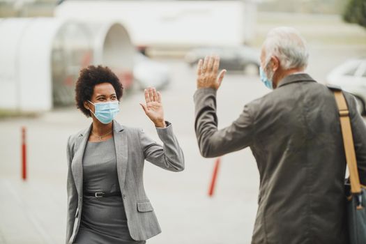 Shot of successful black businesswoman and her senior male colleague using protective mask and greeting with wave hands during COVID-19 pandemic to avoid handshakes.