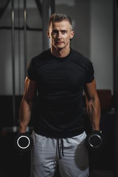 Shot of a muscular guy in sportswear working out at the hard training in the gym. He is pumping up biceps muscule with heavy weight.