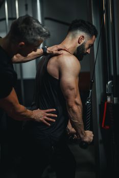 Shot of a muscular guy in sportswear working out with personal trainer at the gym. He is doing cable triceps pulldowns during a strength training.