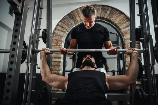 Shot of a muscular guy in sportswear working out with personal trainer at the gym. He is pumping up chest muscule during a hard ttraining.