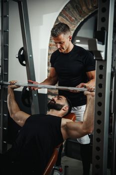 Shot of a muscular guy in sportswear working out with personal trainer at the gym. He is pumping up chest muscule during a hard ttraining.