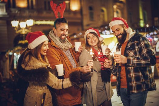 Shot of a group of cheerful young friends having fun with sparklers  and enjoying hot drink on the Christmas market at an evening party.