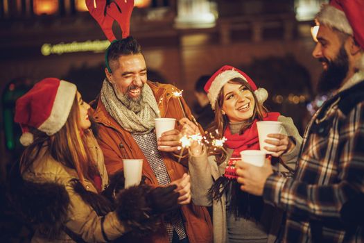 Shot of a group of cheerful young friends having fun with sparklers  and enjoying hot drink on the Christmas market at an evening party.