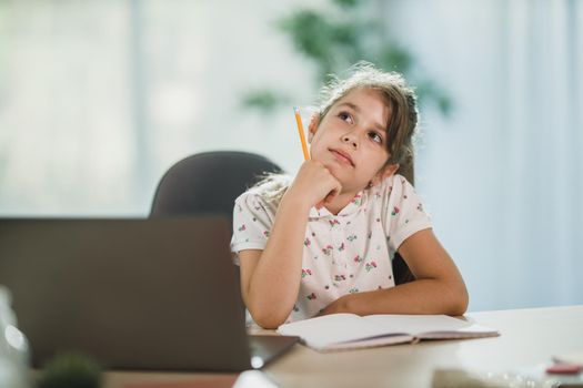 A cute pensive little girl using her laptop and doing a homework assignment at home.