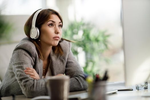 A worried young woman sitting alone in her home office with headphones and working on computer.