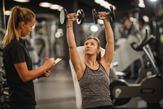 Shot of a muscular young woman in sportswear working out with female personal trainer at the gym. She is pumping up her muscule with dumbbell.