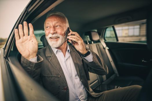 Shot of a successful senior man talking on a smartphone and waving while sitting in the backseat of a car during his business commute.