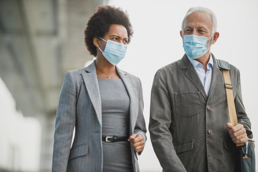 Shot of two successful multi-ethnic business people with protective mask having a discussion while walking in front of the office building during COVID-19 pandemic.