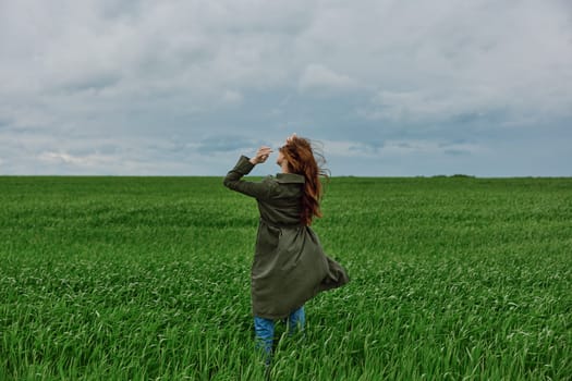 a red-haired woman in a long raincoat stands in a green field in cloudy weather with her back to the camera enjoying nature. High quality photo