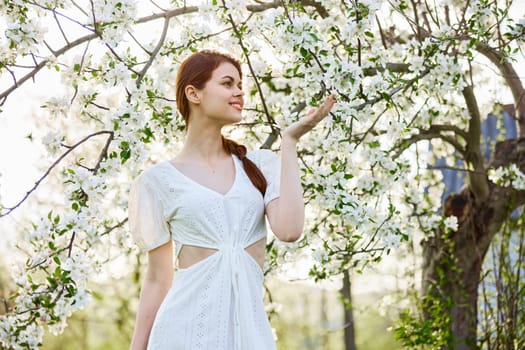 a slender, happy woman in a light dress poses next to a flowering tree in the countryside. High quality photo