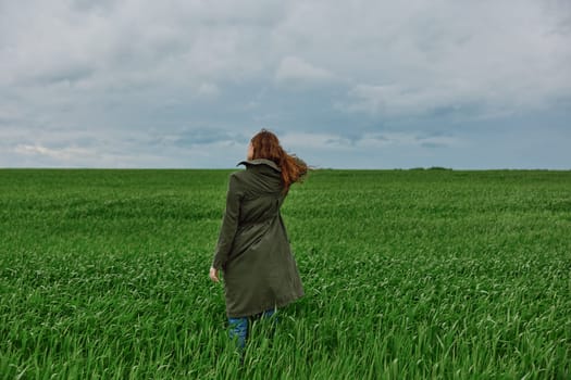 a red-haired woman in a long raincoat stands in a green field in cloudy weather with her back to the camera enjoying nature. High quality photo