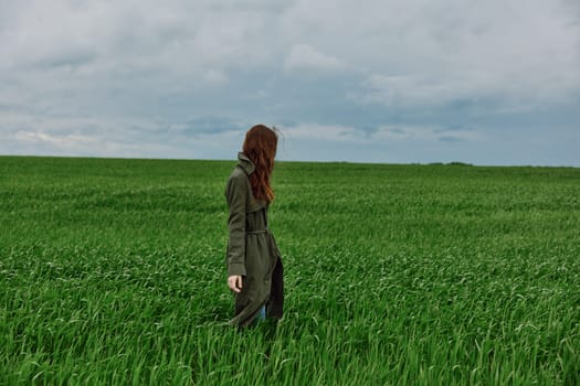 a red-haired woman in a long raincoat stands in a green field in cloudy weather with her back to the camera enjoying nature. High quality photo