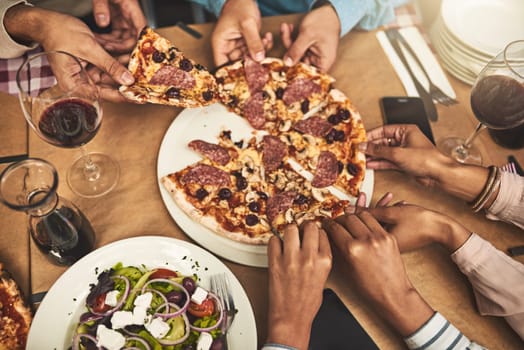 Dig in. High angle shot of a group of unrecognizable peoples hands each grabbing a slice of pizza while being seated at a restaurant