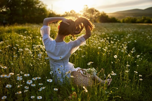 a red-haired woman in a light dress sits with her back to the camera in a field of daisies straightening her hair with her hands against the backdrop of the setting sun. High quality photo