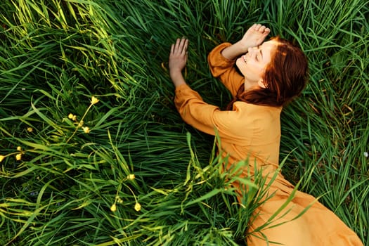 a relaxed red-haired woman enjoys summer lying in the tall green grass in a long orange dress smiling happily looking away. High quality photo