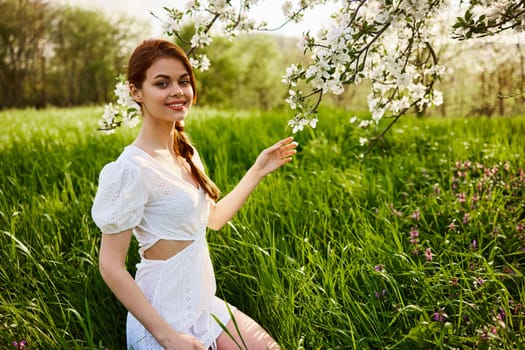 a lovely woman in a light short dress sits near a flowering tree and smiles while looking at the camera. High quality photo
