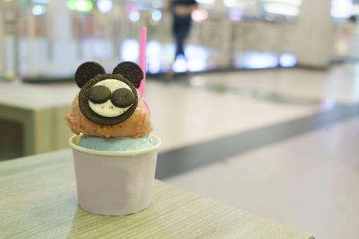A cup of gelato with mouse-like biscuit topping served on the wooden table with blurred background.