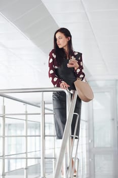 Holding phone. Beautiful young brunette in black skirt indoors in office or airport. Having free time.