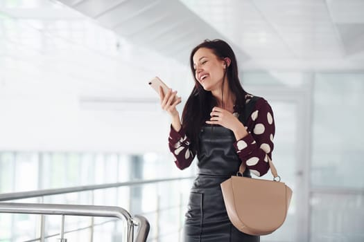 Holding phone. Beautiful young brunette in black skirt indoors in office or airport. Having free time.