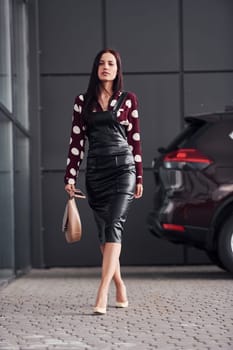 Young brunette in black skirt and with bag in hands standing against black automobile.