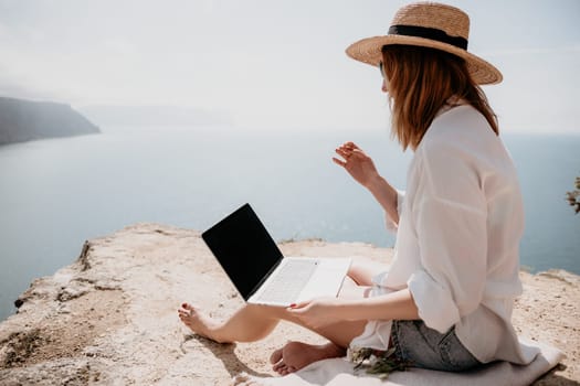 Successful business woman in yellow hat working on laptop by the sea. Pretty lady typing on computer at summer day outdoors. Freelance, travel and holidays concept.