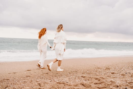 Women sea walk friendship spring. Two girlfriends, redhead and blonde, middle-aged walk along the sandy beach of the sea, dressed in white clothes. Against the backdrop of a cloudy sky and the winter sea. Weekend concept