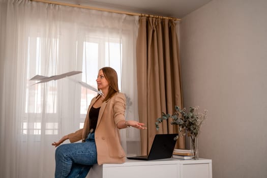 Professional woman sitting on desk in home office, positive female lawyer rejoices throwing papers up. She is wearing a beige jacket and jeans.