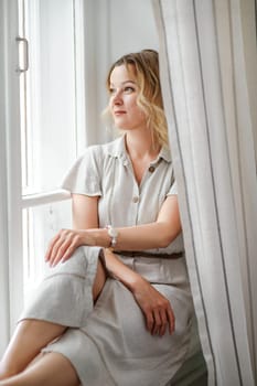 A middle-aged woman in a cream dress sits mysteriously and looks out the window on the windowsill. Green trees outside