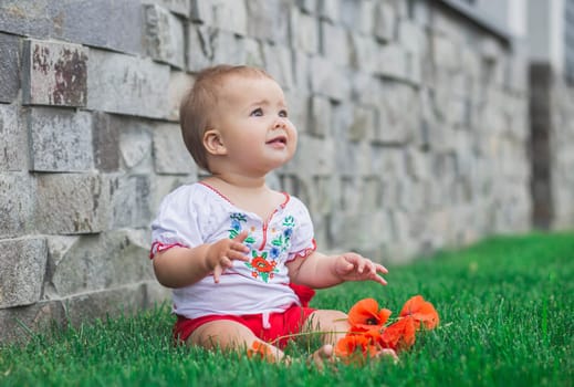 baby in embroidered shirt sitting on a lawn with flowers.