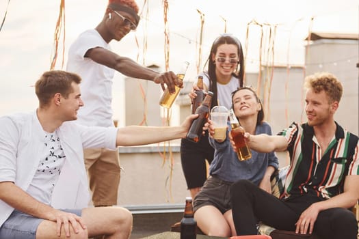 Doing cheers by bottles with beer. Group of young people in casual clothes have a party at rooftop together at daytime.