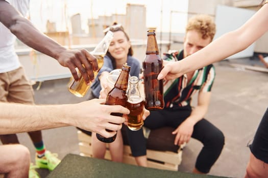 Doing cheers by bottles with beer. Group of young people in casual clothes have a party at rooftop together at daytime.