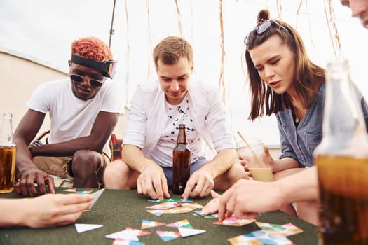 Playing card game. Group of young people in casual clothes have a party at rooftop together at daytime.