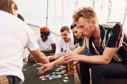 Playing card game. Group of young people in casual clothes have a party at rooftop together at daytime.