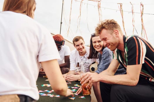 Playing card game. Group of young people in casual clothes have a party at rooftop together at daytime.