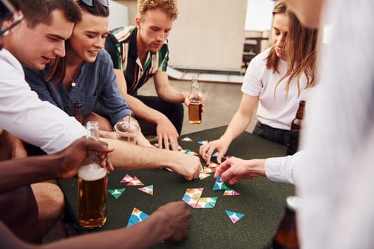 Playing card game. Group of young people in casual clothes have a party at rooftop together at daytime.