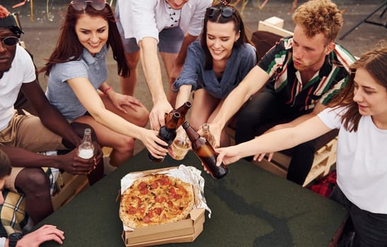 Doing cheers by bottles with beer. Group of young people in casual clothes have a party at rooftop together at daytime.