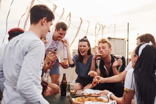 With delicious pizza. Group of young people in casual clothes have a party at rooftop together at daytime.