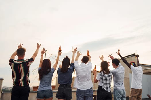 Standing high and looking at the cloudy sky. Group of young people in casual clothes have a party at rooftop together at daytime.