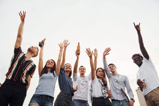 Standing high and looking at the cloudy sky. Group of young people in casual clothes have a party at rooftop together at daytime.