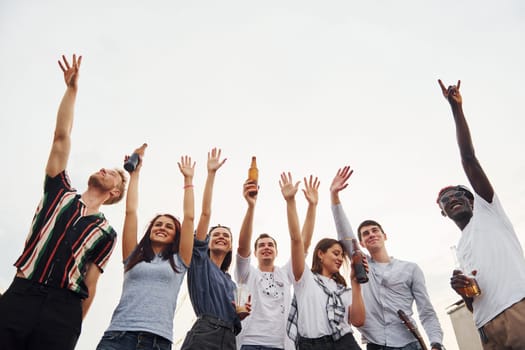 Standing high and looking at the cloudy sky. Group of young people in casual clothes have a party at rooftop together at daytime.