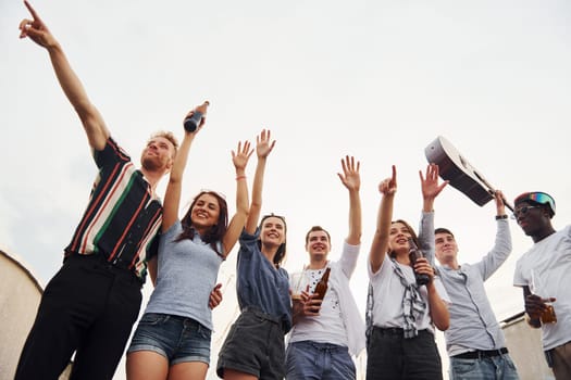 Standing high and looking at the cloudy sky. Group of young people in casual clothes have a party at rooftop together at daytime.