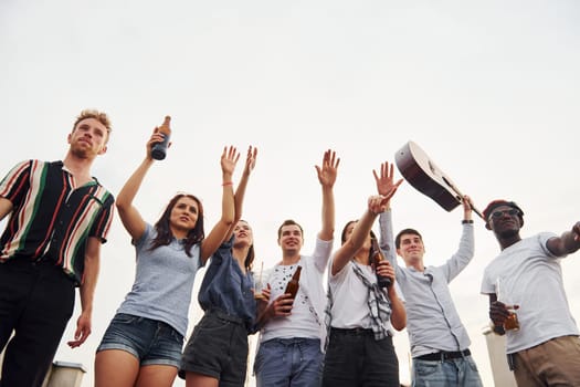 Standing high and looking at the cloudy sky. Group of young people in casual clothes have a party at rooftop together at daytime.