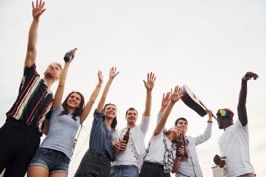 Standing high and looking at the cloudy sky. Group of young people in casual clothes have a party at rooftop together at daytime.