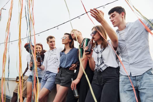 Leaning on the edge of the rooftop with decorates. Group of young people in casual clothes have a party together at daytime.